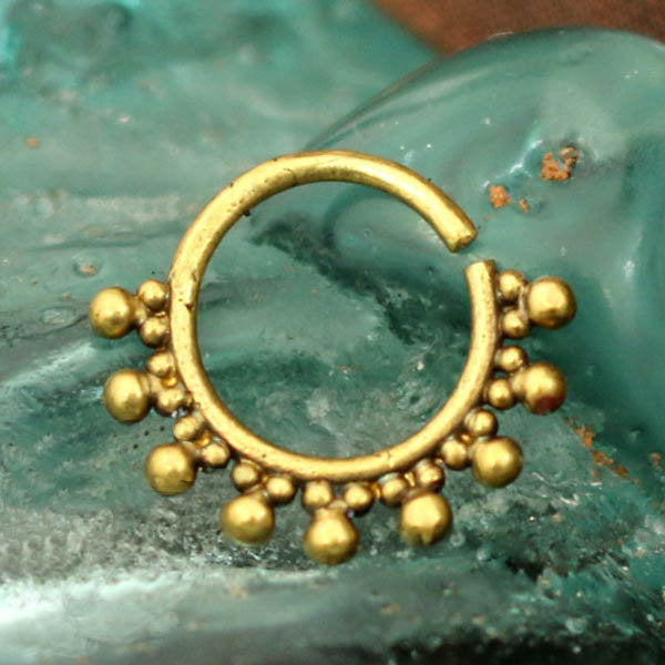 The BRAI Seamless Septum Ring in Gold, crafted by Kaya Movement, boasts a circular gold brass design adorned with small gold beads neatly arranged around the lower half. The backdrop is a textured, teal-colored surface. This ring's tribal ethnic and ornate design offers versatility as a multi-use piercing accessory.